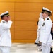 Two U.S. Navy officers in white dress uniforms are exchanging salutes during what appears to be a formal military ceremony. A male officer on the left and a female officer on the right face each other mid-salute, with the female officer displaying multiple service ribbons/medals on her uniform. Seated civilians and a third uniformed individual are visible in the background.