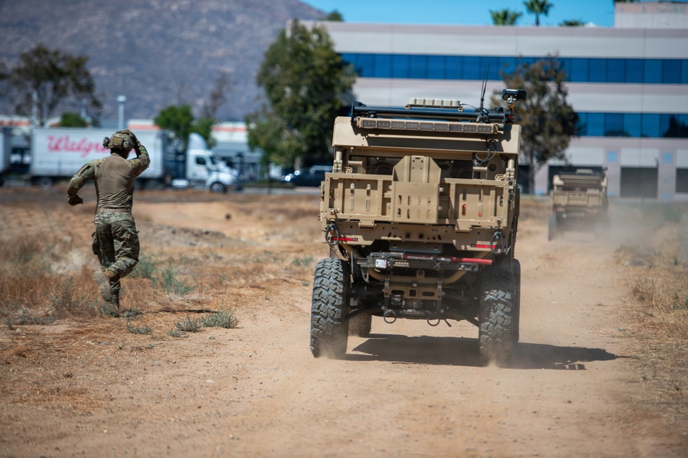 A military personnel in camouflage uniform is running on a dirt path alongside a tan/desert-colored military tactical vehicle, with a second vehicle visible in the background. The scene appears to take place in a semi-urban area near commercial buildings and a Walgreens distribution truck. The setting suggests a military training exercise or operational movement in a Southern California-type environment based on the terrain and vegetation.