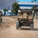 A military personnel in camouflage uniform is running on a dirt path alongside a tan/desert-colored military tactical vehicle, with a second vehicle visible in the background. The scene appears to take place in a semi-urban area near commercial buildings and a Walgreens distribution truck. The setting suggests a military training exercise or operational movement in a Southern California-type environment based on the terrain and vegetation.