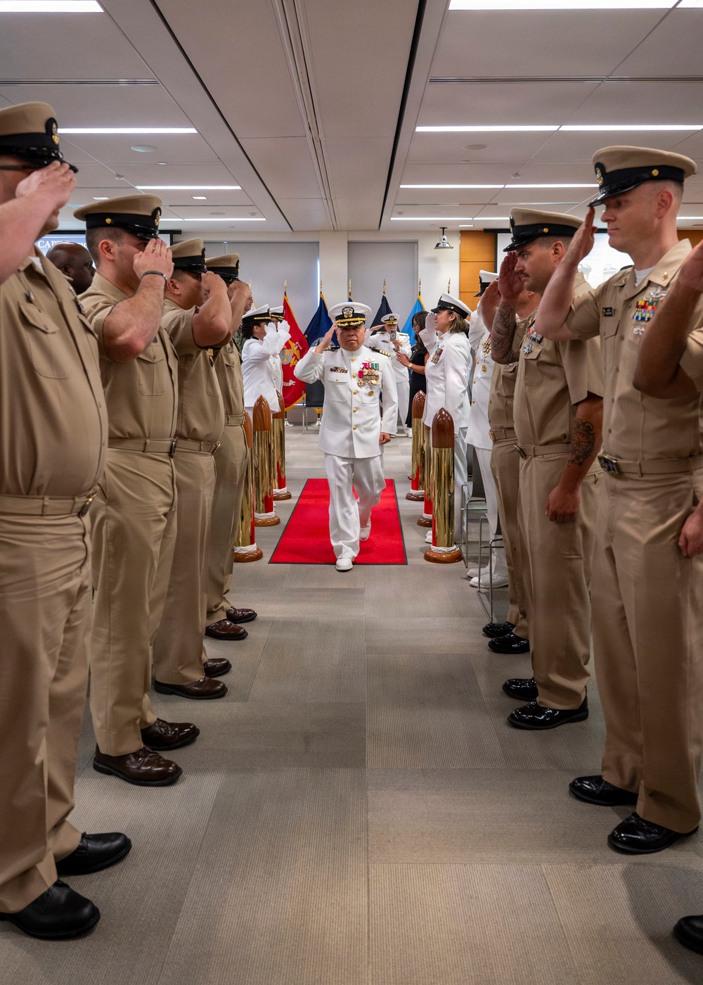 A U.S. Navy officer in white dress uniform walks down a red carpet through a sideboy honor corridor while personnel in tan/khaki uniforms render salutes on both sides. The ceremony appears to be a formal military event such as a change of command, retirement, or official arrival/departure. Brass shell casing stanchions line the red carpet, and military flags are visible in the background.