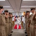 A U.S. Navy officer in white dress uniform walks down a red carpet through a sideboy honor corridor while personnel in tan/khaki uniforms render salutes on both sides. The ceremony appears to be a formal military event such as a change of command, retirement, or official arrival/departure. Brass shell casing stanchions line the red carpet, and military flags are visible in the background.