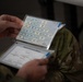 A person wearing military camouflage (OCP/MultiCam pattern) is holding open a laminated reference booklet or flip-card set. The visible pages show a chemical flowchart/reaction diagram and a page titled 'Chemical Structures (Blister)' with molecular structure drawings. This appears to be a military CBRN (Chemical, Biological, Radiological, Nuclear) training reference material.