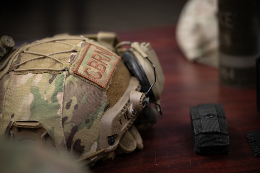 A military tactical helmet covered in MultiCam/OCP camouflage pattern fabric is displayed on a wooden surface. The helmet features a 'CBRN' (Chemical, Biological, Radiological, and Nuclear) patch, rail-mounted ear protection, and elastic retention netting. Additional military gear including a black MOLLE pouch is visible beside the helmet.