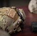 A military tactical helmet covered in MultiCam/OCP camouflage pattern fabric is displayed on a wooden surface. The helmet features a 'CBRN' (Chemical, Biological, Radiological, and Nuclear) patch, rail-mounted ear protection, and elastic retention netting. Additional military gear including a black MOLLE pouch is visible beside the helmet.