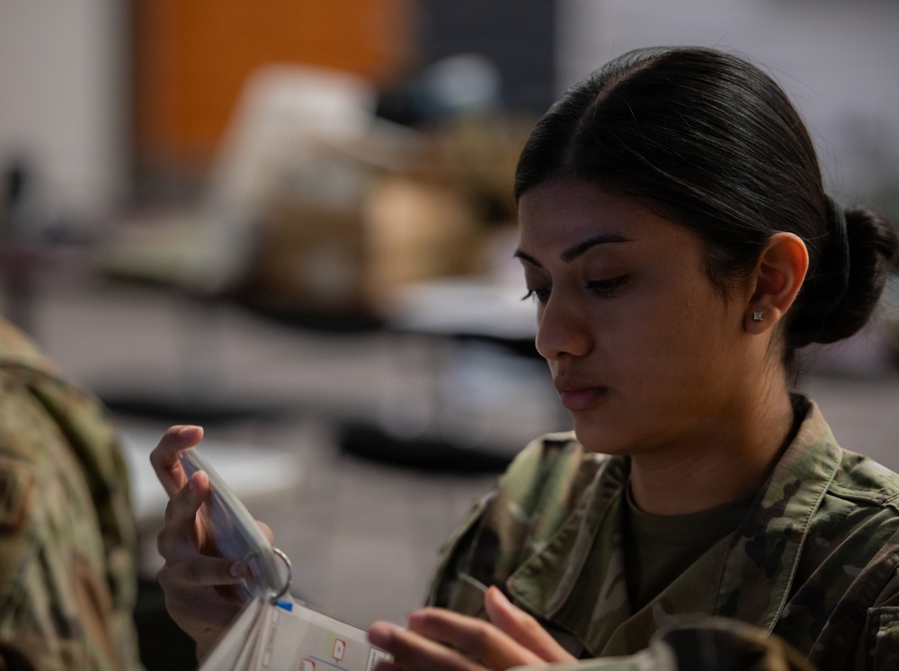 A female military service member in camouflage uniform is closely examining a small transparent object, possibly a medical or equipment item. She appears focused and engaged in a detailed inspection or preparation task. The setting appears to be an indoor military facility or classroom environment.
