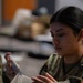 A female military service member in camouflage uniform is closely examining a small transparent object, possibly a medical or equipment item. She appears focused and engaged in a detailed inspection or preparation task. The setting appears to be an indoor military facility or classroom environment.