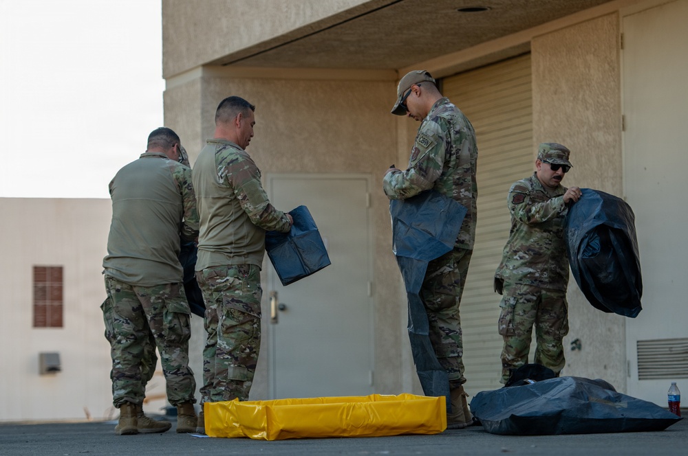 Four military personnel in OCP (Operational Camouflage Pattern) uniforms are handling what appear to be equipment bags or covers outside a building. They are working together near a door and garage-style roll-up door on a paved surface. A yellow containment berm or barrier is visible on the ground alongside dark-colored bags or equipment covers.