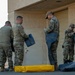 Four military personnel in OCP (Operational Camouflage Pattern) uniforms are handling what appear to be equipment bags or covers outside a building. They are working together near a door and garage-style roll-up door on a paved surface. A yellow containment berm or barrier is visible on the ground alongside dark-colored bags or equipment covers.
