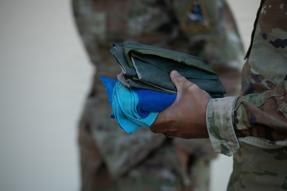 A close-up photograph showing the hand of a military personnel in OCP (Operational Camouflage Pattern) uniform holding a folded olive-drab/green fabric item along with blue microfiber cleaning cloths. A second individual in the same camouflage uniform is visible in the blurred background. The focus is on the items being held, suggesting a maintenance, cleaning, or equipment distribution activity.