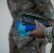 A close-up photograph showing the hand of a military personnel in OCP (Operational Camouflage Pattern) uniform holding a folded olive-drab/green fabric item along with blue microfiber cleaning cloths. A second individual in the same camouflage uniform is visible in the blurred background. The focus is on the items being held, suggesting a maintenance, cleaning, or equipment distribution activity.