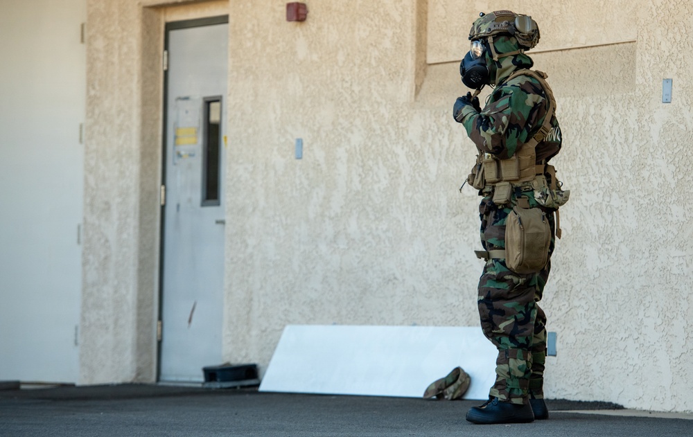 A military or law enforcement personnel is standing outside a building wearing full CBRN (Chemical, Biological, Radiological, Nuclear) protective gear including a gas mask, tactical helmet, camouflage uniform, and tactical vest. The individual appears to be in a stationary position near a building entrance, possibly during a training exercise or operational scenario. Additional gear is visible on the ground nearby.
