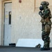 A military or law enforcement personnel is standing outside a building wearing full CBRN (Chemical, Biological, Radiological, Nuclear) protective gear including a gas mask, tactical helmet, camouflage uniform, and tactical vest. The individual appears to be in a stationary position near a building entrance, possibly during a training exercise or operational scenario. Additional gear is visible on the ground nearby.