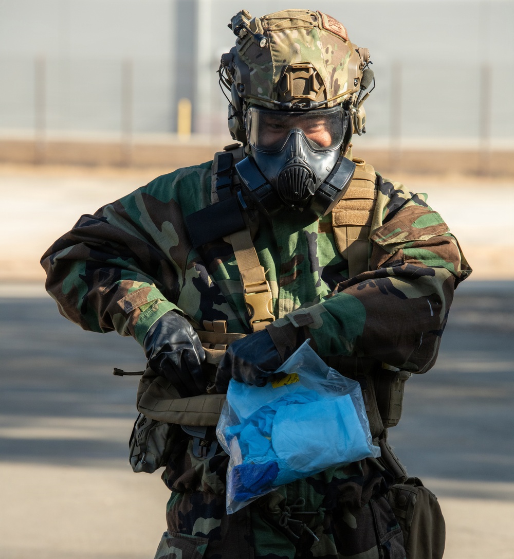 A military personnel wearing full CBRN (Chemical, Biological, Radiological, Nuclear) protective gear is handling a sealed clear plastic bag containing blue materials. The individual is dressed in woodland camouflage BDU, tactical vest, protective gas mask, ballistic helmet with accessories, and black rubber gloves. The setting appears to be an outdoor paved area, likely a military installation.