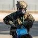 A military personnel wearing full CBRN (Chemical, Biological, Radiological, Nuclear) protective gear is handling a sealed clear plastic bag containing blue materials. The individual is dressed in woodland camouflage BDU, tactical vest, protective gas mask, ballistic helmet with accessories, and black rubber gloves. The setting appears to be an outdoor paved area, likely a military installation.