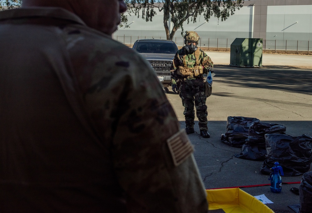 Two military personnel are visible in an outdoor setting, with one wearing full CBRN (Chemical, Biological, Radiological, Nuclear) protective gear including a gas mask, combat helmet, camouflage suit, tactical vest, and blue gloves. The scene appears to be a decontamination or CBRN training exercise, evidenced by a yellow containment tray, black bags, and spray bottles on the ground. A US flag patch is visible on the foreground soldier's uniform sleeve.