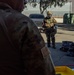Two military personnel are visible in an outdoor setting, with one wearing full CBRN (Chemical, Biological, Radiological, Nuclear) protective gear including a gas mask, combat helmet, camouflage suit, tactical vest, and blue gloves. The scene appears to be a decontamination or CBRN training exercise, evidenced by a yellow containment tray, black bags, and spray bottles on the ground. A US flag patch is visible on the foreground soldier's uniform sleeve.