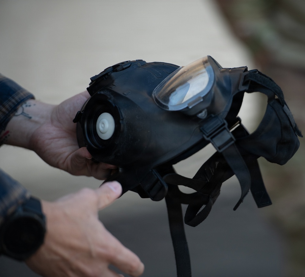A person is holding a black rubber gas mask, displaying it toward the camera. The mask features a single large visor lens, an exhale valve, adjustable head straps, and filter canister port. The individual wearing a plaid/flannel shirt has visible tattoos on their hand and wrist.