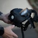 A person is holding a black rubber gas mask, displaying it toward the camera. The mask features a single large visor lens, an exhale valve, adjustable head straps, and filter canister port. The individual wearing a plaid/flannel shirt has visible tattoos on their hand and wrist.