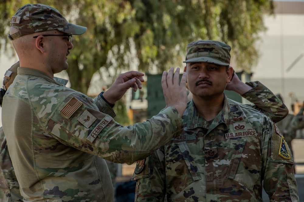 Two U.S. Air Force personnel in OCP (Operational Camouflage Pattern) uniforms are engaged in what appears to be a promotion or award pinning ceremony outdoors. The individual on the left, identified by name tape as 'FEENSTRA,' is reaching toward the cap or collar area of the individual on the right, identified as 'KAASI,' suggesting a rank insignia is being pinned or adjusted. The setting appears to be a military installation with trees and equipment visible in the background.