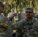 Two U.S. Air Force personnel in OCP (Operational Camouflage Pattern) uniforms are engaged in what appears to be a promotion or award pinning ceremony outdoors. The individual on the left, identified by name tape as 'FEENSTRA,' is reaching toward the cap or collar area of the individual on the right, identified as 'KAASI,' suggesting a rank insignia is being pinned or adjusted. The setting appears to be a military installation with trees and equipment visible in the background.