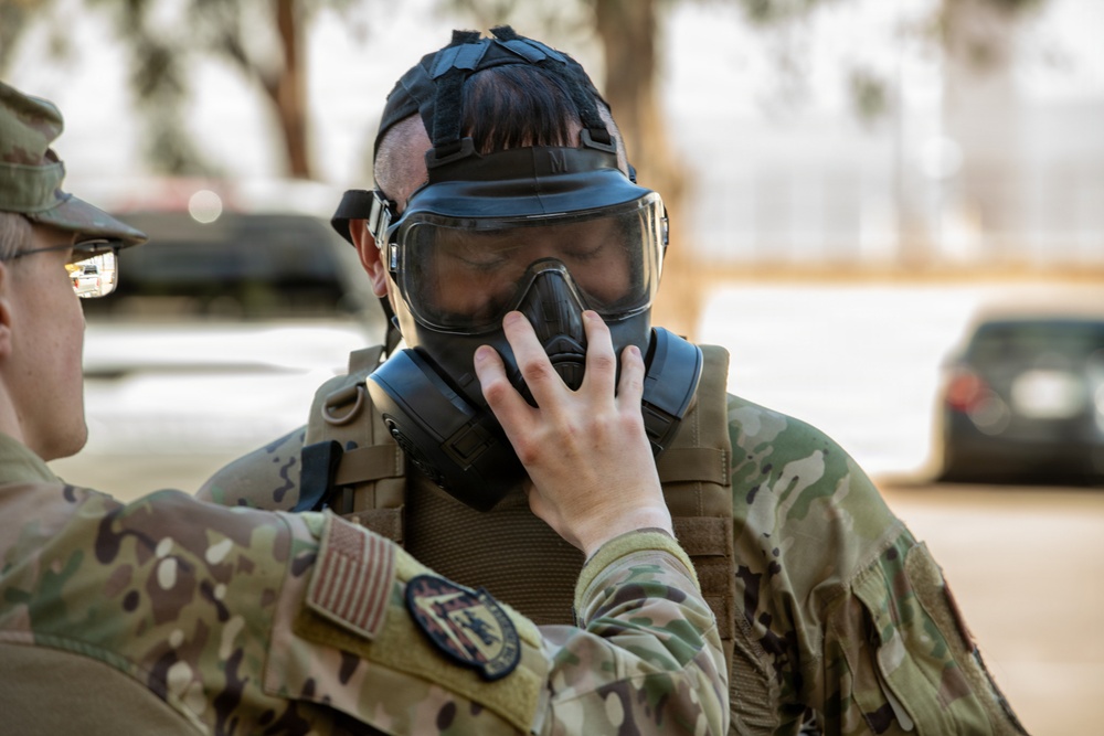Two military personnel in MultiCam/OCP camouflage uniforms are engaged in what appears to be a gas mask fitting or inspection exercise. One soldier is assisting the other in properly seating or checking the seal of a military gas mask. The setting appears to be an outdoor area on a military installation.