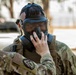 Two military personnel in MultiCam/OCP camouflage uniforms are engaged in what appears to be a gas mask fitting or inspection exercise. One soldier is assisting the other in properly seating or checking the seal of a military gas mask. The setting appears to be an outdoor area on a military installation.