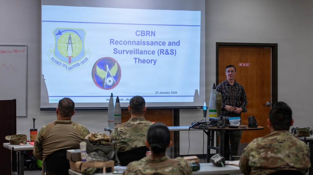 A civilian instructor is delivering a CBRN (Chemical, Biological, Radiological, and Nuclear) Reconnaissance and Surveillance Theory class to a group of military personnel in uniform. The presentation slide, dated 23 January 2026, displays the Air Force Civil Engineer Center logo. Several ordnance or training munition models are visible on a table near the presenter.