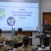 A civilian instructor is delivering a CBRN (Chemical, Biological, Radiological, and Nuclear) Reconnaissance and Surveillance Theory class to a group of military personnel in uniform. The presentation slide, dated 23 January 2026, displays the Air Force Civil Engineer Center logo. Several ordnance or training munition models are visible on a table near the presenter.
