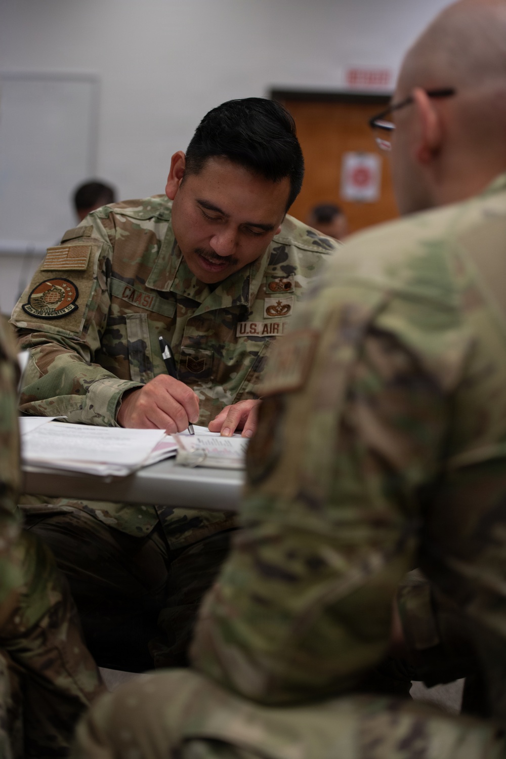 A U.S. Air Force service member, identified by the name tape 'CANASI' and 'U.S. AIR F[ORCE]' tape, is seated at a desk writing or reviewing documents in what appears to be a classroom or training environment. He is wearing OCP (Operational Camouflage Pattern) uniform with a unit patch on the left shoulder and skill badges on the chest. A second service member in OCP uniform is visible in the foreground, out of focus.