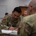 A U.S. Air Force service member, identified by the name tape 'CANASI' and 'U.S. AIR F[ORCE]' tape, is seated at a desk writing or reviewing documents in what appears to be a classroom or training environment. He is wearing OCP (Operational Camouflage Pattern) uniform with a unit patch on the left shoulder and skill badges on the chest. A second service member in OCP uniform is visible in the foreground, out of focus.