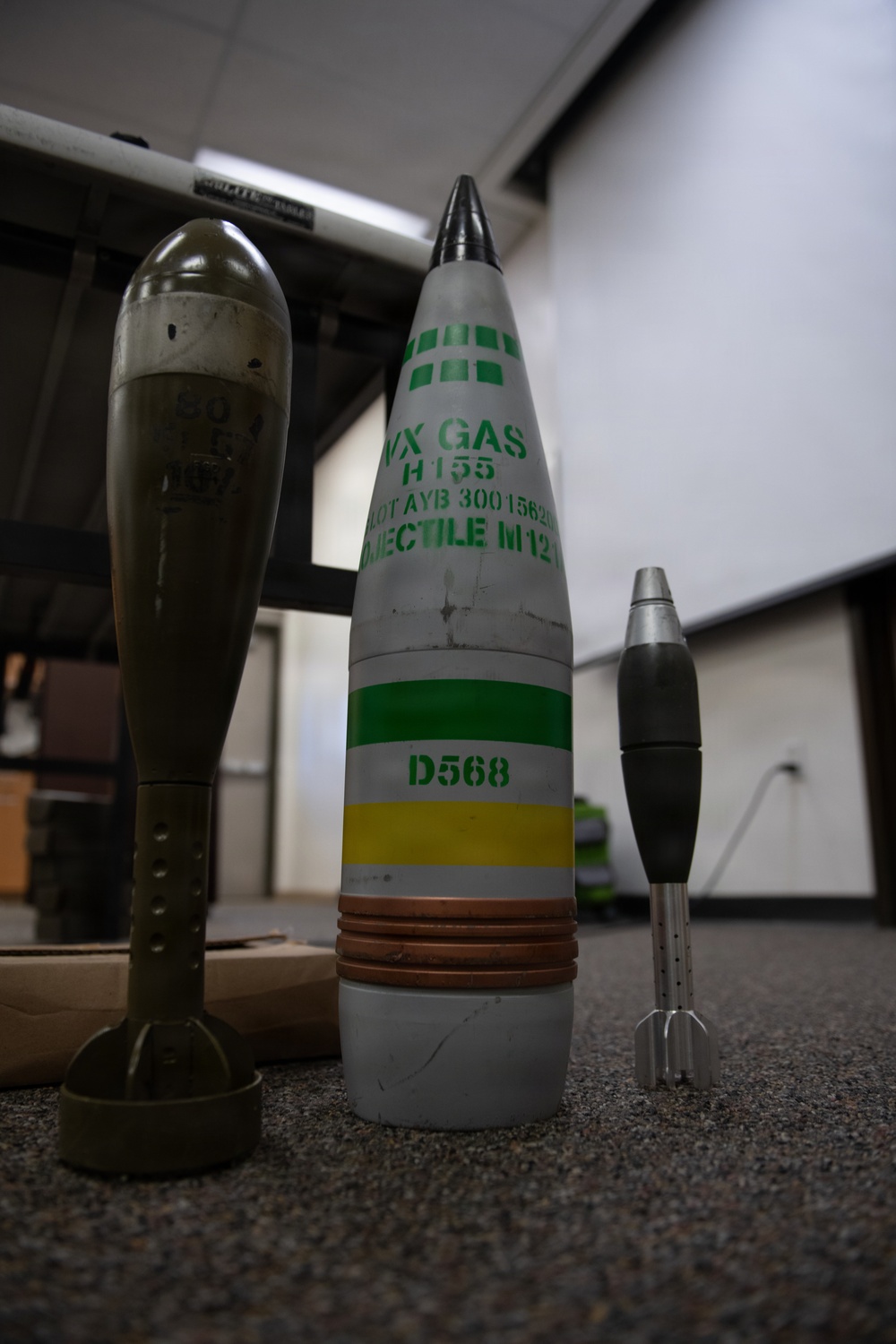 The image shows three munition training/display items arranged on a carpeted floor in what appears to be an indoor classroom or briefing room setting. The central item is a large artillery projectile labeled 'VX GAS H155' with markings 'PROJECTILE M121', lot number 'AYB 300156', and color-coded bands (green, yellow) with identifier 'D568'. These appear to be inert training or display rounds used for chemical weapons familiarization or demilitarization training.