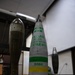 The image shows three munition training/display items arranged on a carpeted floor in what appears to be an indoor classroom or briefing room setting. The central item is a large artillery projectile labeled 'VX GAS H155' with markings 'PROJECTILE M121', lot number 'AYB 300156', and color-coded bands (green, yellow) with identifier 'D568'. These appear to be inert training or display rounds used for chemical weapons familiarization or demilitarization training.