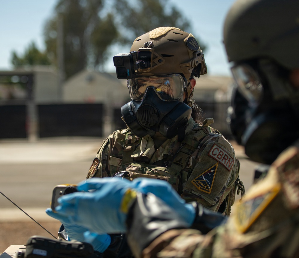 A military personnel wearing full CBRN (Chemical, Biological, Radiological, and Nuclear) protective gear is engaged in what appears to be a training exercise or operational scenario. The individual is equipped with a tactical helmet, gas mask with protective goggles, and camouflage uniform bearing a CBRN patch and Space Force insignia. A second person wearing blue nitrile gloves is handling equipment in the foreground.
