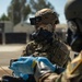 A military personnel wearing full CBRN (Chemical, Biological, Radiological, and Nuclear) protective gear is engaged in what appears to be a training exercise or operational scenario. The individual is equipped with a tactical helmet, gas mask with protective goggles, and camouflage uniform bearing a CBRN patch and Space Force insignia. A second person wearing blue nitrile gloves is handling equipment in the foreground.