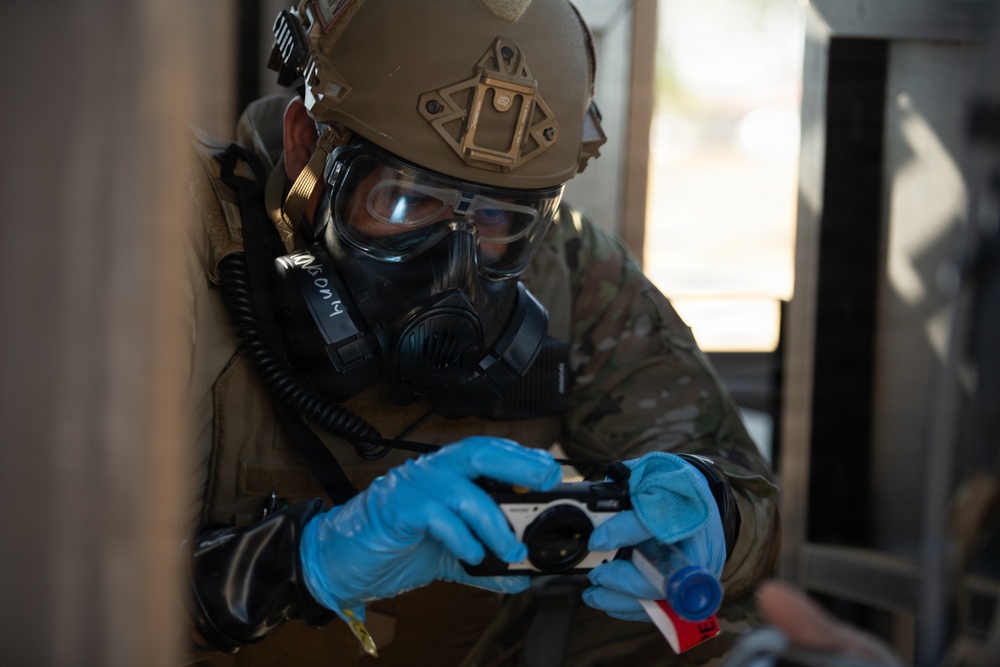 A military or law enforcement personnel wearing full CBRN (Chemical, Biological, Radiological, Nuclear) protective gear is handling what appears to be a detection or sampling device. The individual is equipped with a tactical helmet, gas mask, protective goggles, camouflage uniform, and blue nitrile gloves. The scene suggests a hazardous materials assessment or CBRN training/response exercise.