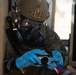 A military or law enforcement personnel wearing full CBRN (Chemical, Biological, Radiological, Nuclear) protective gear is handling what appears to be a detection or sampling device. The individual is equipped with a tactical helmet, gas mask, protective goggles, camouflage uniform, and blue nitrile gloves. The scene suggests a hazardous materials assessment or CBRN training/response exercise.