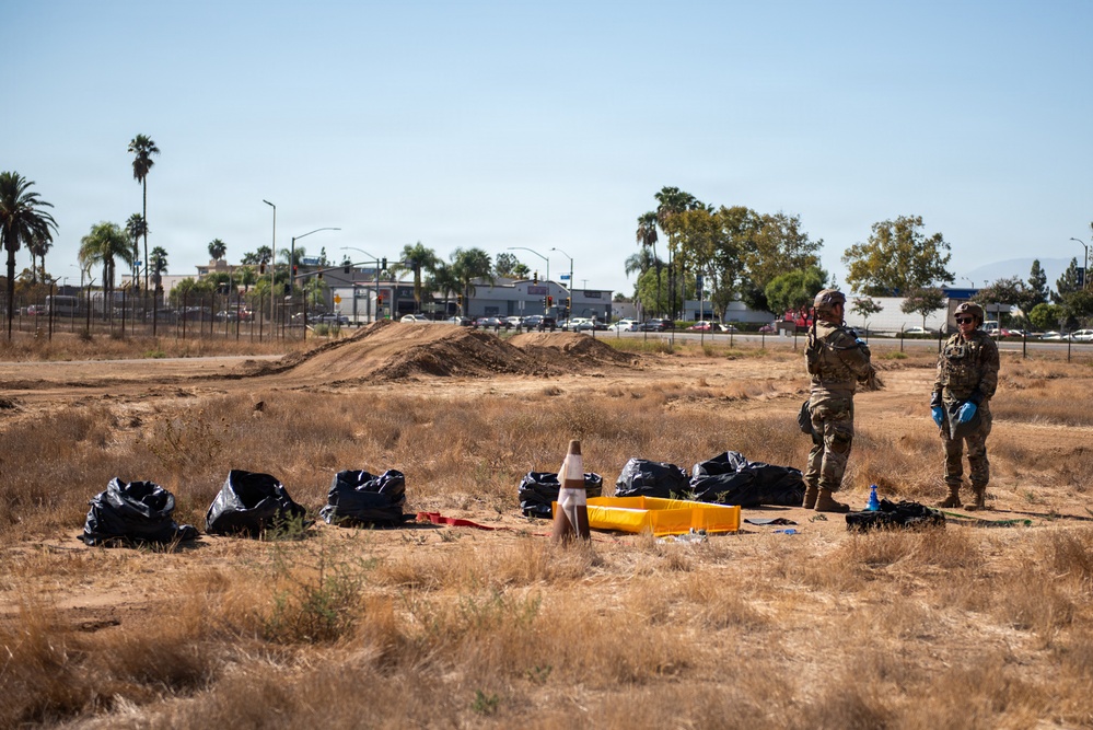 Two individuals in military/tactical camouflage uniforms wearing blue gloves stand in a dry, open field surrounded by multiple black garbage bags and yellow containers. The scene appears to depict a cleanup or collection operation in an arid urban-adjacent area. Palm trees, commercial buildings, and a busy road are visible in the background, suggesting a Southern California or similar warm-climate urban setting.