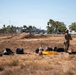 Two individuals in military/tactical camouflage uniforms wearing blue gloves stand in a dry, open field surrounded by multiple black garbage bags and yellow containers. The scene appears to depict a cleanup or collection operation in an arid urban-adjacent area. Palm trees, commercial buildings, and a busy road are visible in the background, suggesting a Southern California or similar warm-climate urban setting.