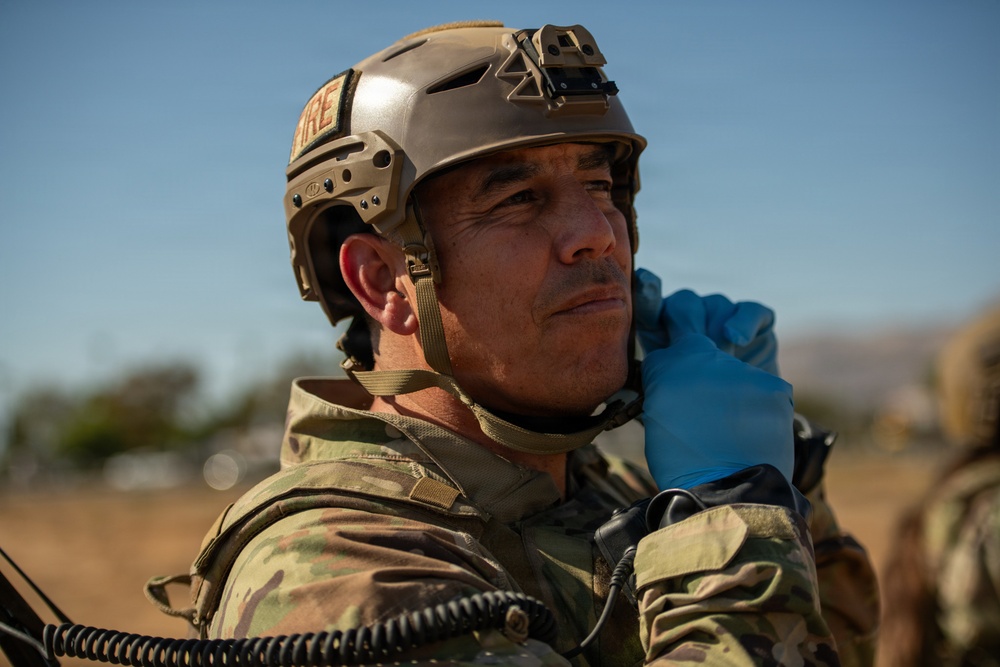 A military personnel member wearing tactical gear is photographed in an outdoor field environment. The individual is wearing a tan/coyote tactical helmet with a mounted device, multicam camouflage uniform, and blue nitrile gloves, and appears to be adjusting communications equipment at their neck. The setting appears to be an arid or semi-arid training or operational environment under clear blue skies.