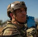 A military personnel member wearing tactical gear is photographed in an outdoor field environment. The individual is wearing a tan/coyote tactical helmet with a mounted device, multicam camouflage uniform, and blue nitrile gloves, and appears to be adjusting communications equipment at their neck. The setting appears to be an arid or semi-arid training or operational environment under clear blue skies.