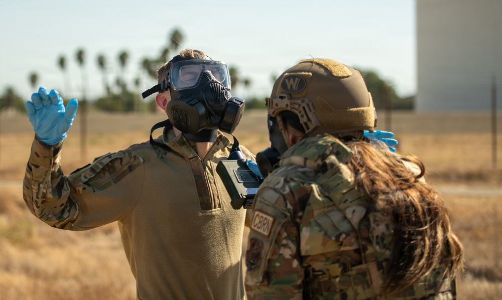 Two military personnel in camouflage uniforms are engaged in what appears to be a CBRN (Chemical, Biological, Radiological, and Nuclear) training exercise or response operation. One individual is wearing a gas mask and blue nitrile gloves with a handheld detection device, while the other wears a tactical helmet marked 'W' and a CBRN patch, also wearing blue nitrile gloves. The setting appears to be an outdoor military or airfield environment with palm trees and dry terrain visible in the background.