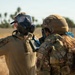 Two military personnel in camouflage uniforms are engaged in what appears to be a CBRN (Chemical, Biological, Radiological, and Nuclear) training exercise or response operation. One individual is wearing a gas mask and blue nitrile gloves with a handheld detection device, while the other wears a tactical helmet marked 'W' and a CBRN patch, also wearing blue nitrile gloves. The setting appears to be an outdoor military or airfield environment with palm trees and dry terrain visible in the background.