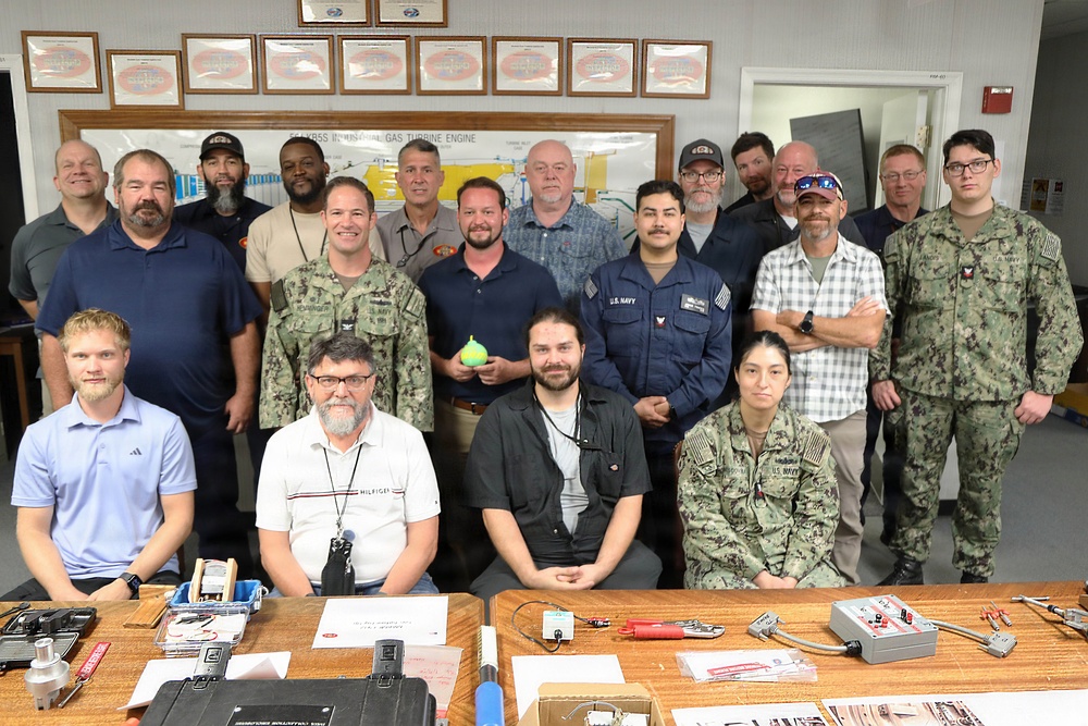 A group of approximately 20 individuals posed for a formal group photo in what appears to be a military training or classroom facility. The group includes both uniformed U.S. Navy personnel in NWU (Navy Working Uniform) camouflage and civilians in casual attire. A workbench in the foreground displays various tools and electronic equipment, suggesting a technical training environment.