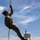 A military or law enforcement personnel in full camouflage uniform is ascending an orange rope using a harness system, suspended in mid-air against a partly cloudy sky. The individual is wearing tactical gear including a helmet, gloves, sunglasses, and combat boots. A water tower and power lines are visible in the background, suggesting a training facility or military base environment.
