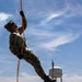 A military or law enforcement personnel in full camouflage uniform is ascending an orange rope using a harness system, suspended in mid-air against a partly cloudy sky. The individual is wearing tactical gear including a helmet, gloves, sunglasses, and combat boots. A water tower and power lines are visible in the background, suggesting a training facility or military base environment.