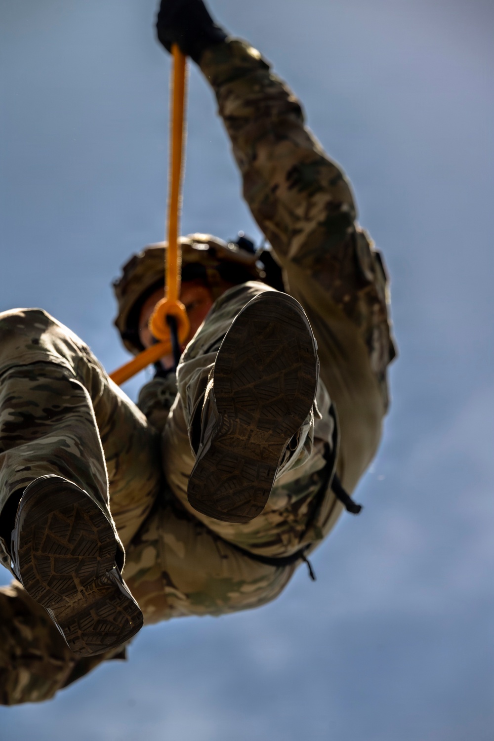A military personnel in full camouflage uniform and tactical helmet is suspended in the air, photographed from a low angle looking upward. The individual is gripping an orange rope or strap while their boots face directly toward the camera. This appears to be a training exercise involving aerial or rope-based operations.