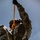 A military personnel in full camouflage uniform and tactical helmet is suspended in the air, photographed from a low angle looking upward. The individual is gripping an orange rope or strap while their boots face directly toward the camera. This appears to be a training exercise involving aerial or rope-based operations.