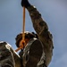 A military personnel in full camouflage uniform and tactical helmet is suspended in the air, photographed from a low angle looking upward. The individual is gripping an orange rope or strap while their boots face directly toward the camera. This appears to be a training exercise involving aerial or rope-based operations.