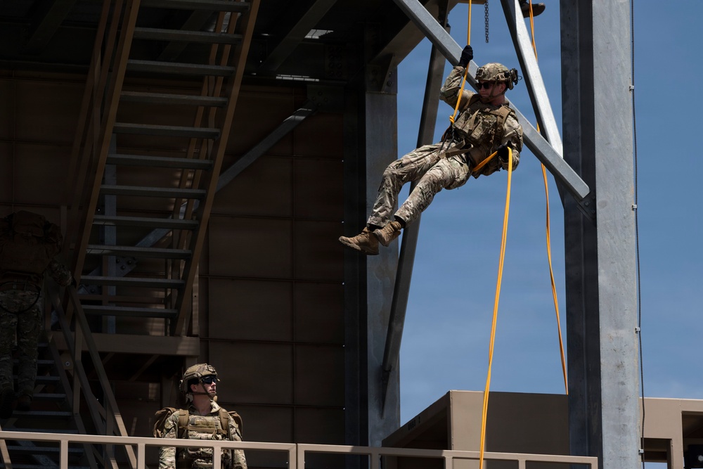 Two military personnel are engaged in a rappelling or rope descent training exercise at what appears to be a dedicated training facility. One soldier is suspended mid-air on orange/yellow ropes attached to a steel tower structure, while a second soldier observes from a platform below. Both are wearing full combat gear including helmets, body armor, and camouflage uniforms.