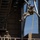 Two military personnel are engaged in a rappelling or rope descent training exercise at what appears to be a dedicated training facility. One soldier is suspended mid-air on orange/yellow ropes attached to a steel tower structure, while a second soldier observes from a platform below. Both are wearing full combat gear including helmets, body armor, and camouflage uniforms.